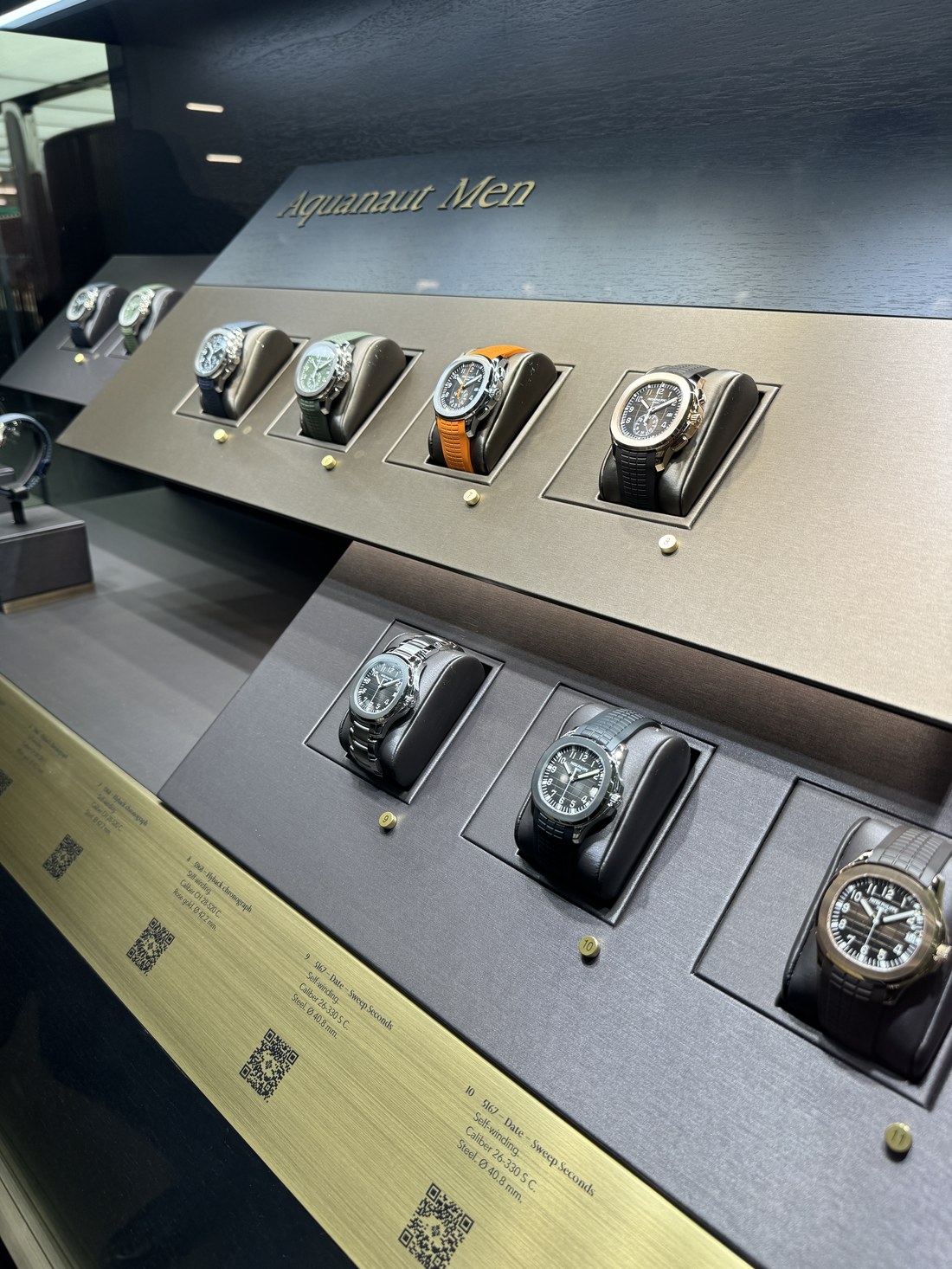 國外錶展現場的展櫃，標示牌刻著 Aquanaut Men，櫃內依序陳列多只不同錶帶與錶盤的腕錶，下方標籤列出型號、機芯與尺寸
